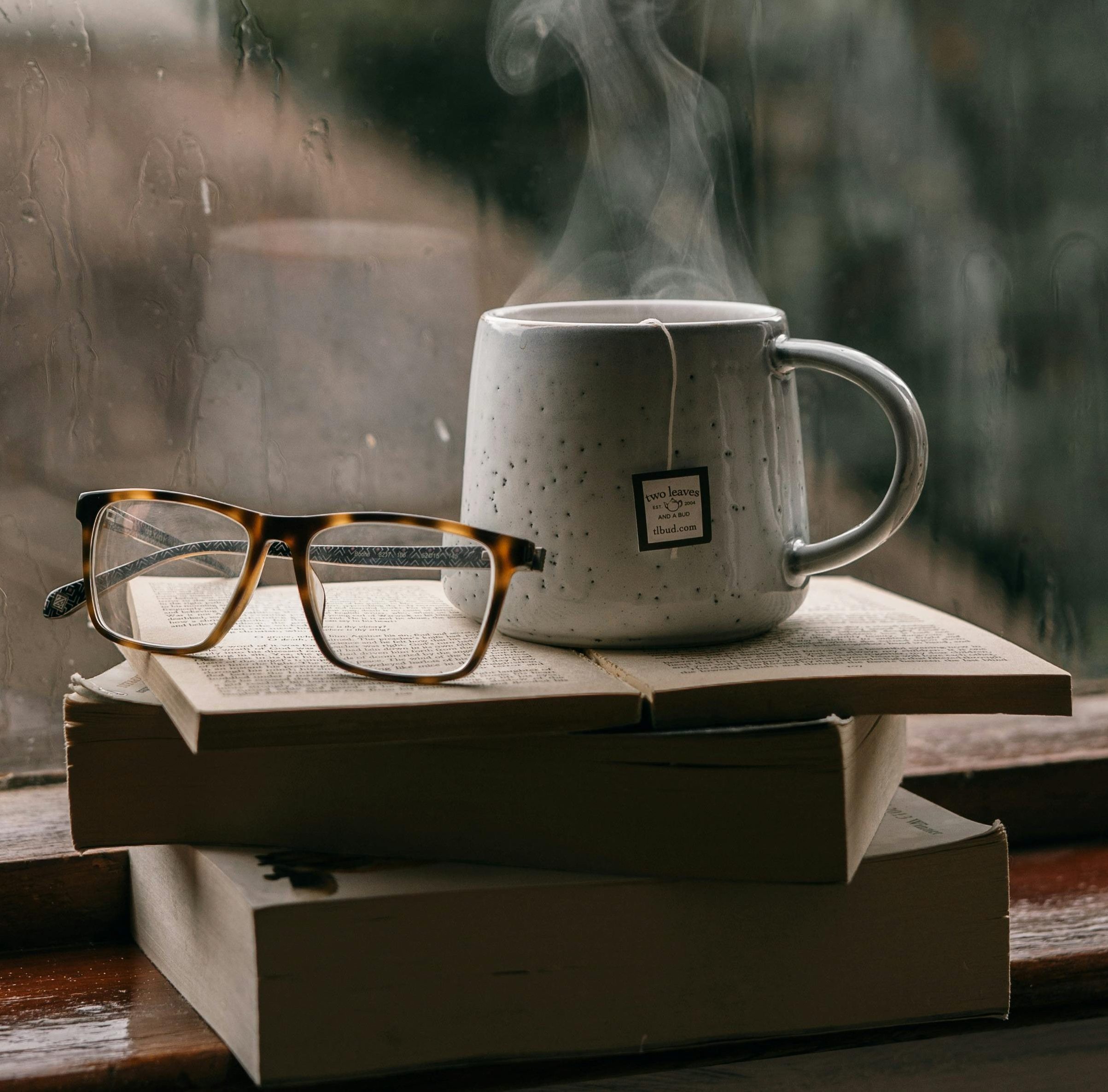 A book and glasses beside a coffee mug on a table by a window, representing reflection, reading, and thoughtful leadership development